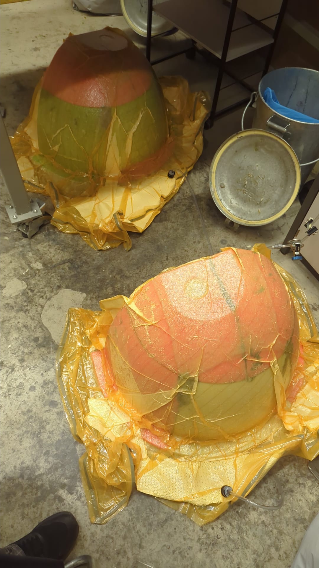 Two eggshells under yellow vacuum bagging film, curing on the workshop floor